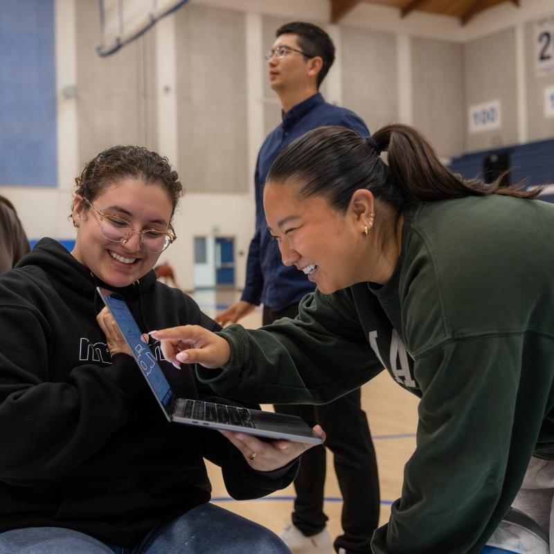 One student smiles and points to the screen of a laptop another student is holding